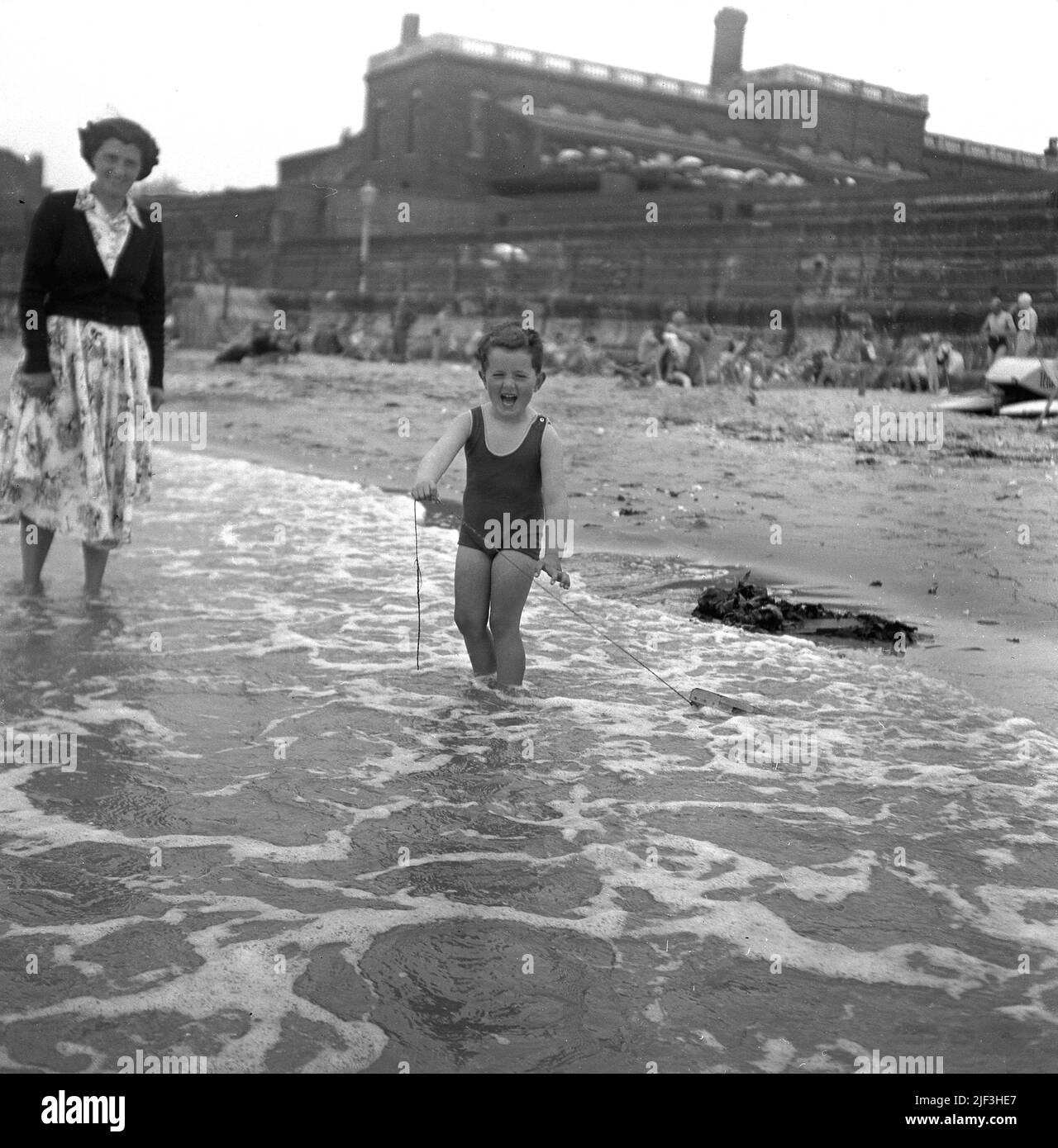 1953, historique, une mère avec son jeune fils dans les eaux peu profondes du bord de mer, avec le petit garçon debout tenant la corde attachée à son bateau en bois jouet qu'il tire à travers l'eau, Margate, Kent, Angleterre, Royaume-Uni. Banque D'Images