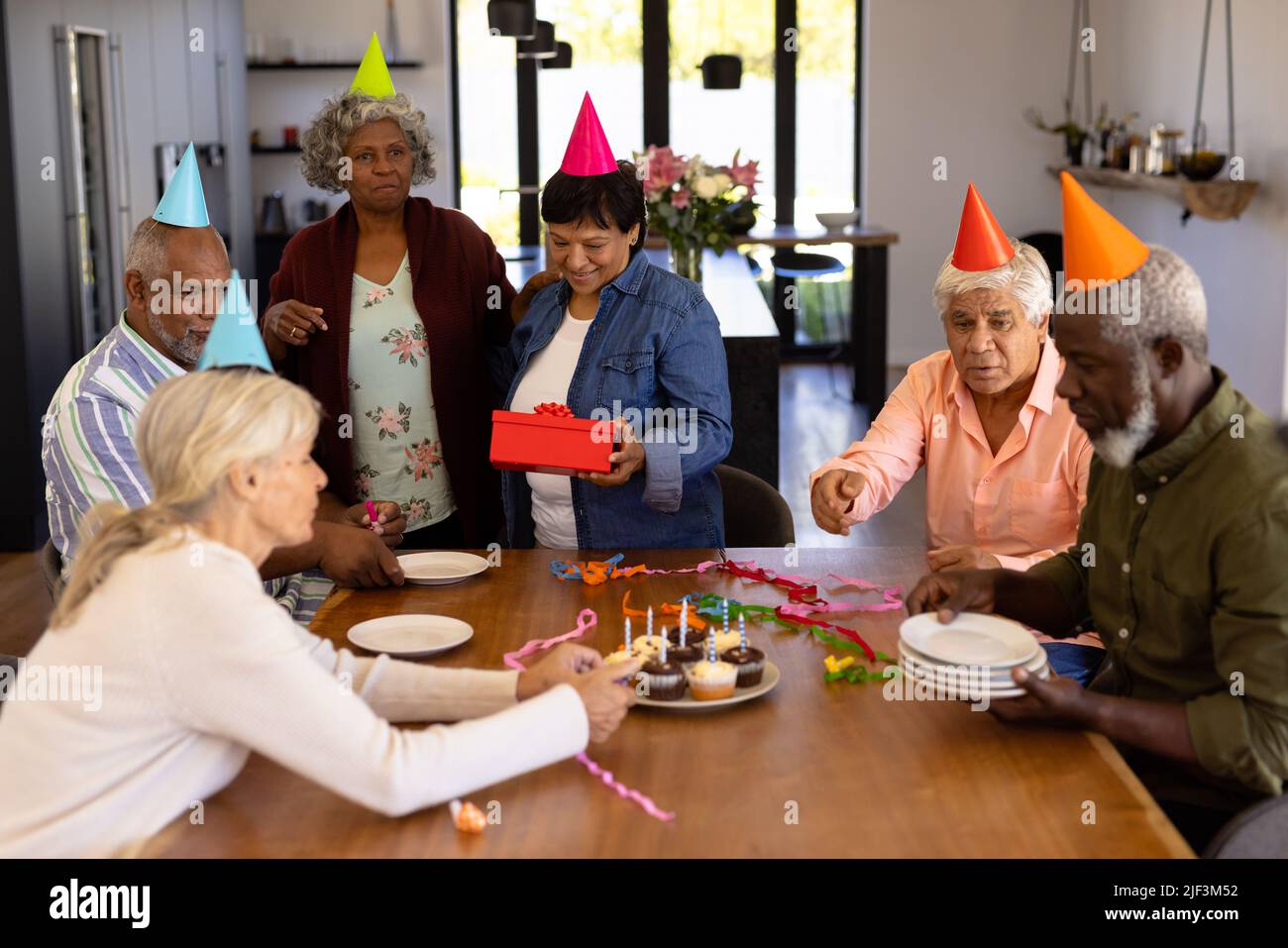 Des amis séniors multiraciaux portant des chapeaux de fête faisant la préparation tandis que la femme tient un cadeau d'anniversaire Banque D'Images