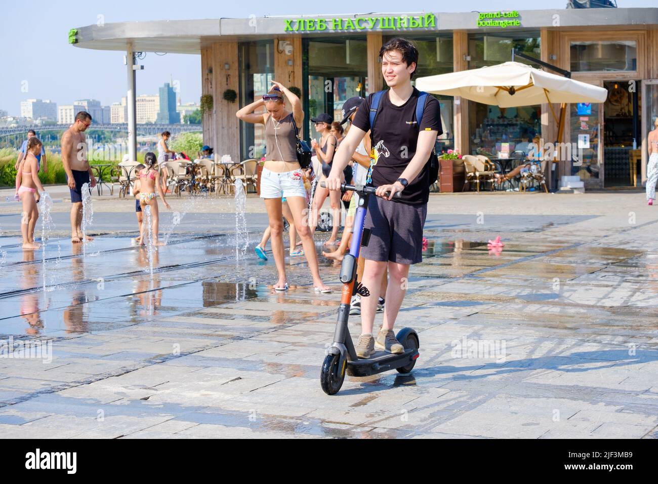 Moscou. Russie. 26 juin 2021. Un jeune homme passe un scooter électrique devant une fontaine où les gens se baignent. Passe-temps amusant lors d'une chaude journée d'été dans la ville. Banque D'Images