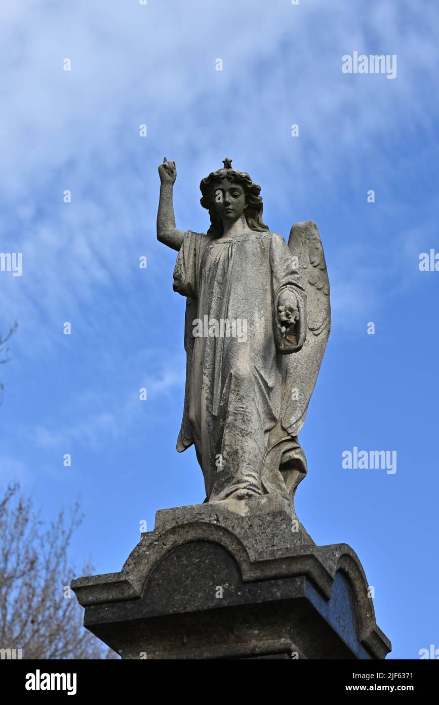 Sculpture en pierre usée d'un ange pointant vers le ciel tout en regardant vers le bas, avec ciel bleu et nuages de lumière dans l'arrière-plan Banque D'Images