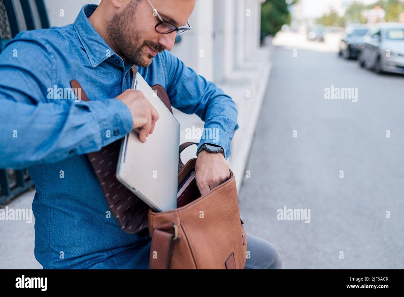 Un jeune homme d'affaires garde son ordinateur portable dans son sac. Homme professionnel de transport dans les coffals est assis contre le bâtiment. Il travaille sur le trottoir dans la ville. Banque D'Images
