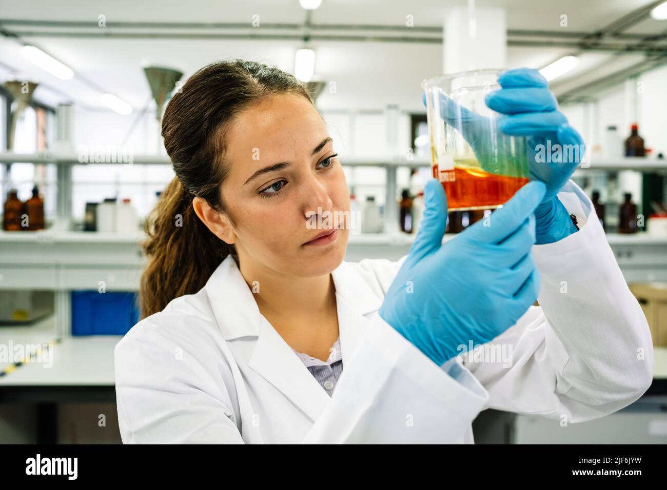 Chercheuse ciblée dans des lunettes de protection et des gants en latex observant un flacon avec un liquide chimique brillant tout en effectuant des recherches en laboratoire léger Banque D'Images