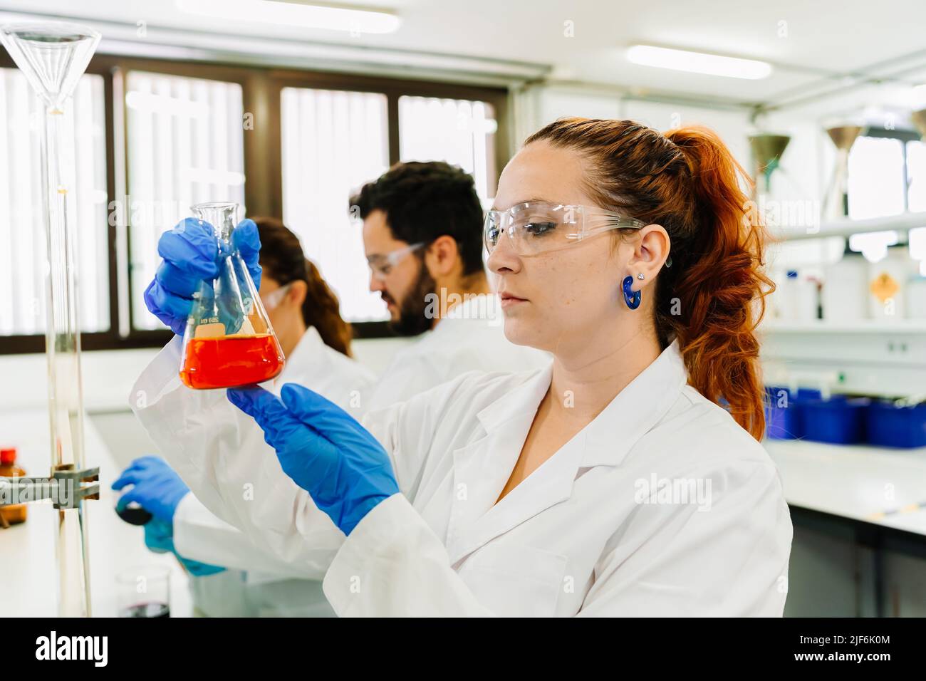 Chercheuse ciblée dans des lunettes de protection et des gants en latex observant un flacon avec un liquide chimique brillant tout en effectuant des recherches en laboratoire léger Banque D'Images