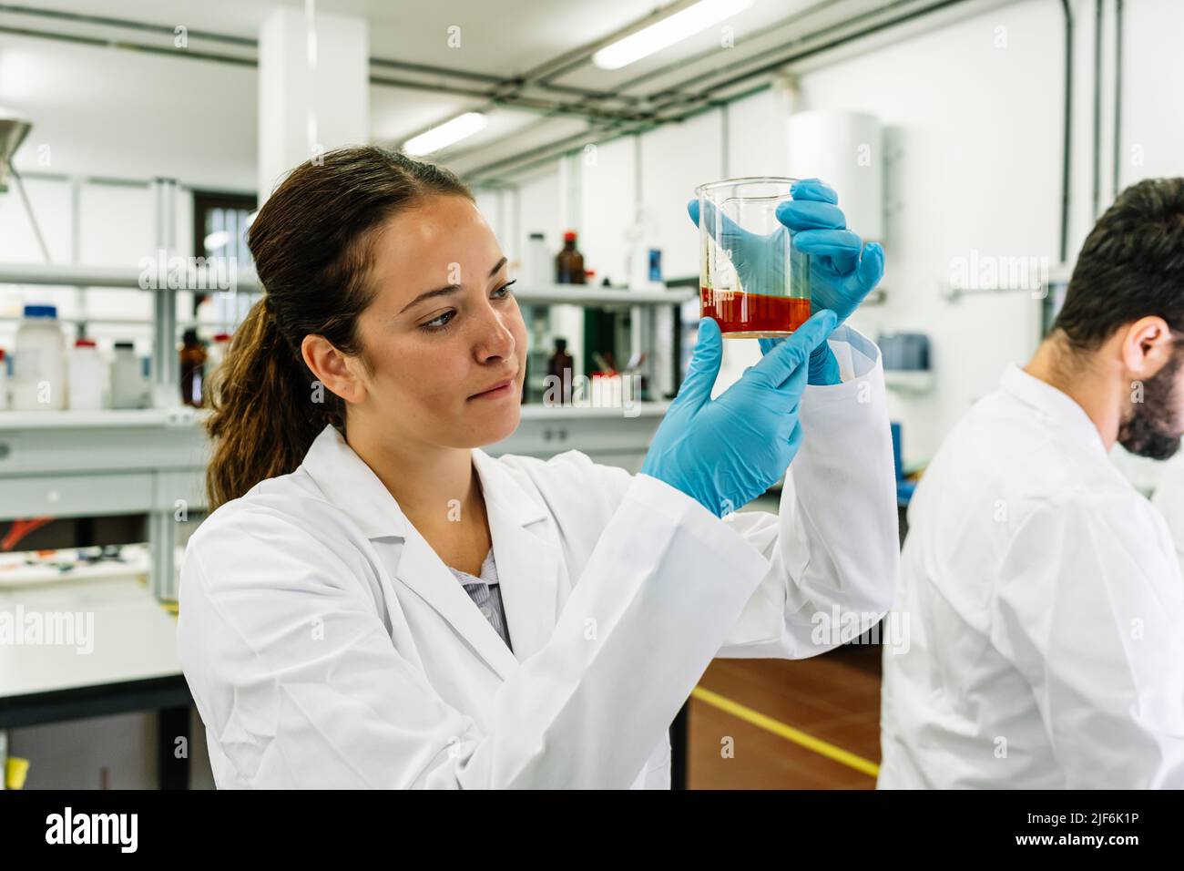 Chercheuse ciblée dans des lunettes de protection et des gants en latex observant un flacon avec un liquide chimique brillant tout en effectuant des recherches en laboratoire léger Banque D'Images