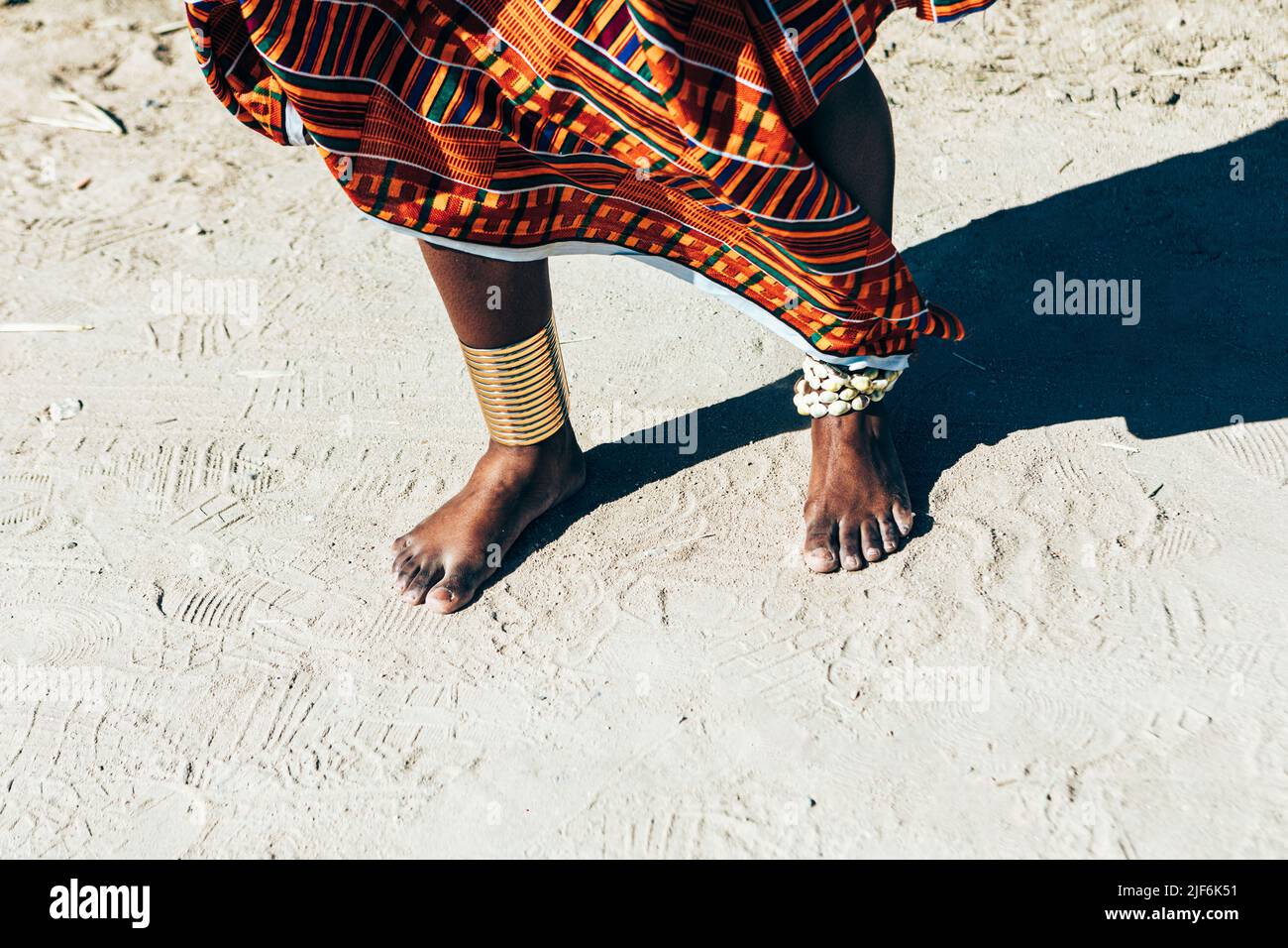 Femelle ethnique non reconnaissable dans un kente tissé à la main coloré portant des anneaux de Kayan sur la cheville tout en se tenant sur la plage de sable Banque D'Images