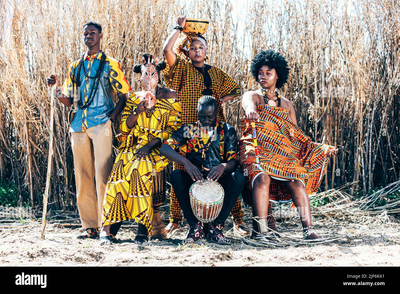Groupe complet de femmes et d'hommes de race noire et mixte dans des tenues africaines authentiques colorées avec des bâtons et des tambours assis et debout contre fie sec Banque D'Images