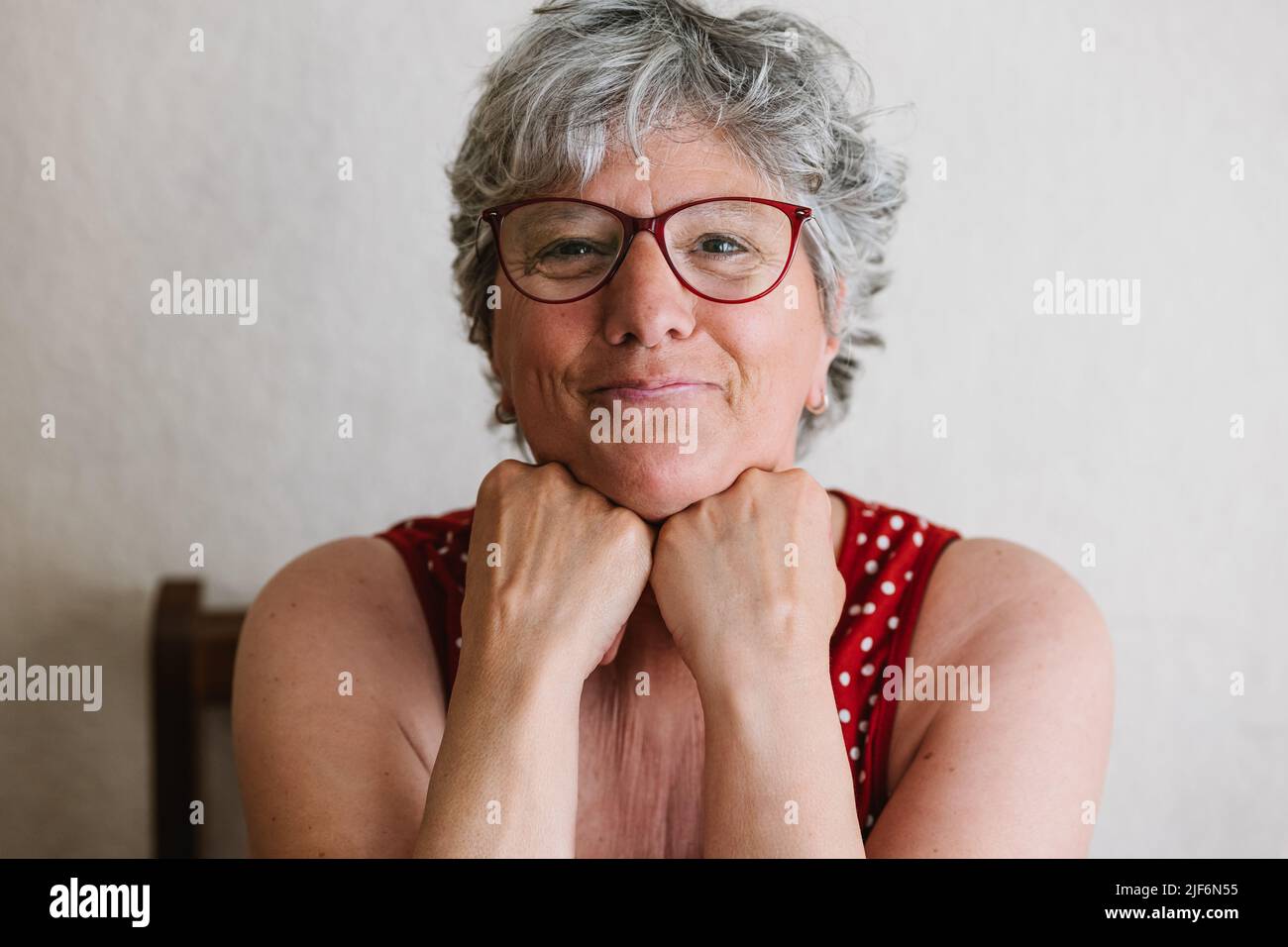 Femme âgée positive avec des cheveux gris dans des vêtements décontractés et des lunettes souriant et regardant l'appareil photo tout en se penchant le menton sur les mains Banque D'Images