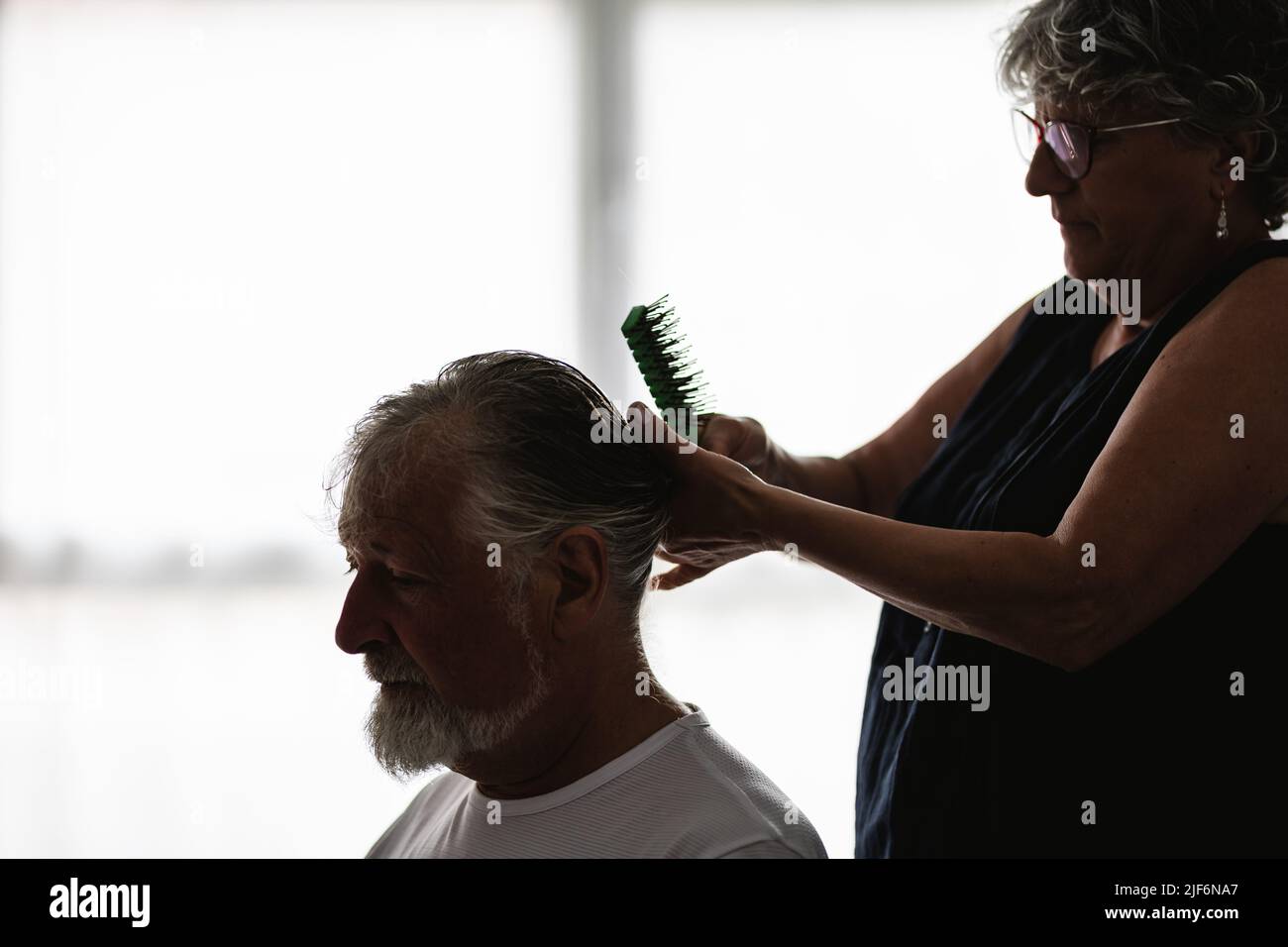 Vue latérale de la petite femme concentrée dans des lunettes avec peigne faisant la coiffure à l'homme âgé barbu dans la pièce lumineuse avec des fenêtres Banque D'Images
