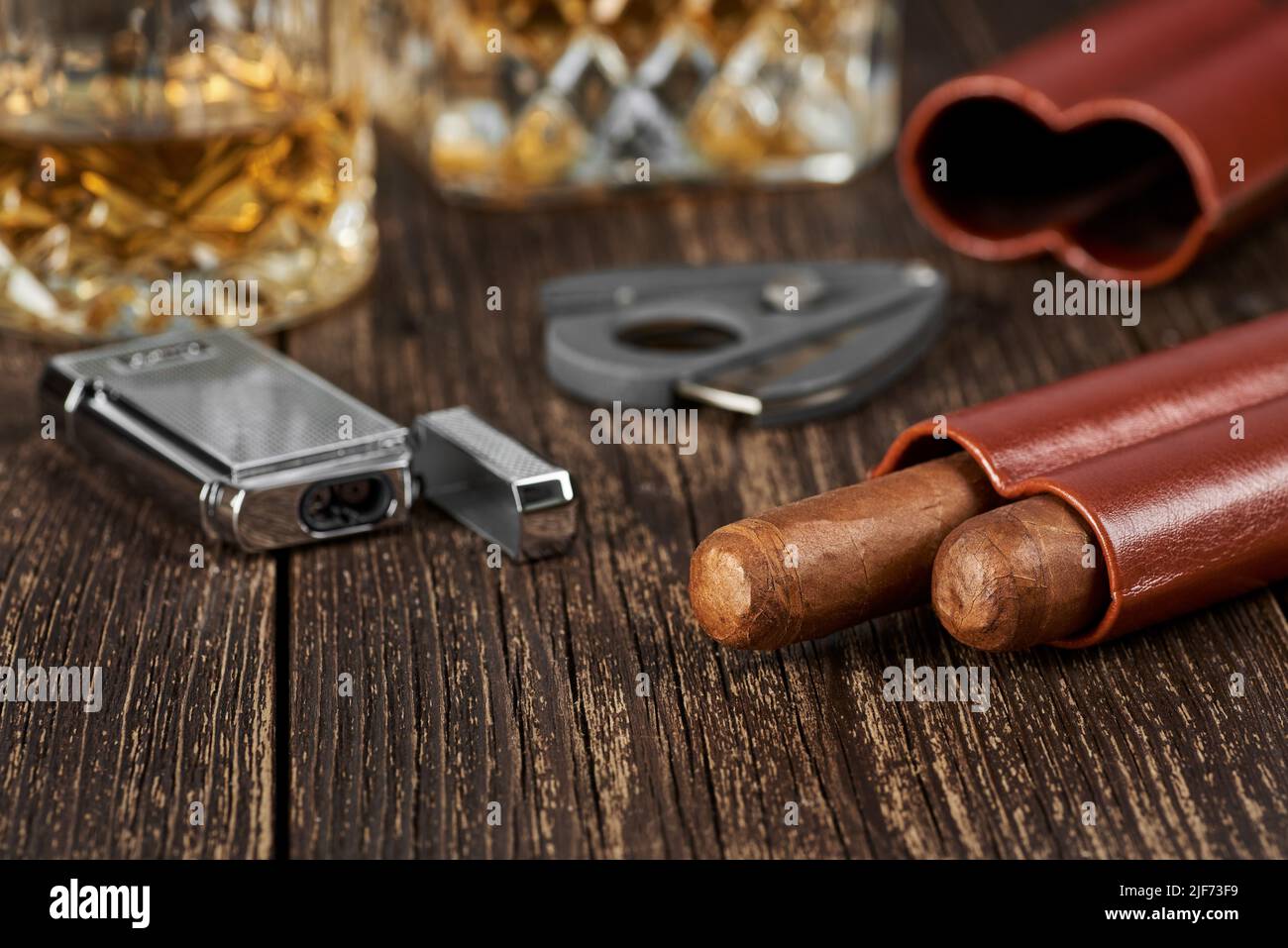 Deux cigares cubains, un couteau et un briquet dans un étui en cuir sur une table brune ancienne. Verre de whisky sur un arrière-plan flou. Banque D'Images