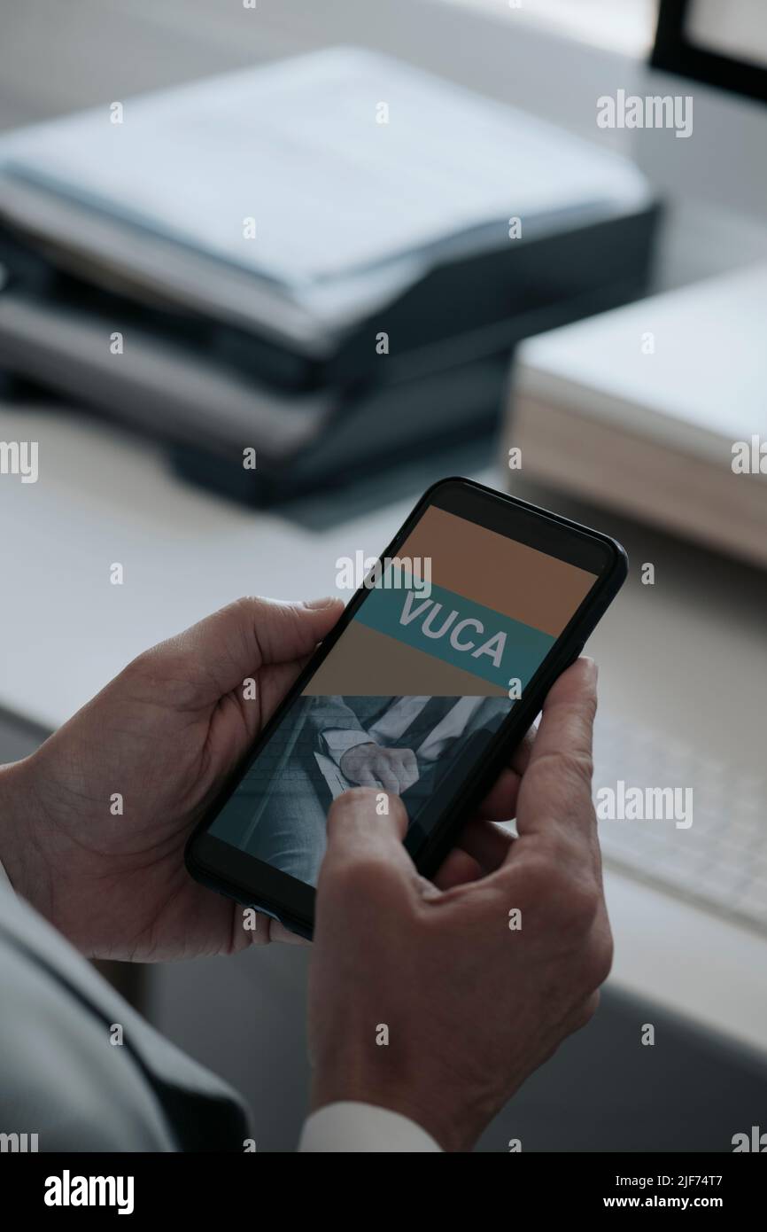 gros plan un homme, portant une chemise blanche et une veste de costume grise, au bureau tenant son smartphone, qui lit le texte VUCA, pour volatilité, incertain Banque D'Images