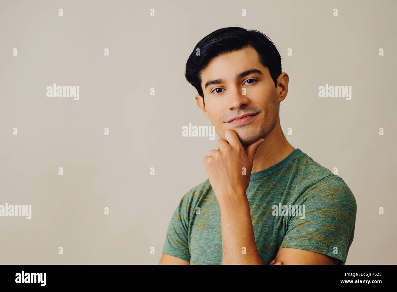 Portrait hispanique latino-homme avec les bras croisés main sur le menton noir cheveux souriant beau jeune adulte vert t-shirt sur fond gris regardant la prise de vue en studio Banque D'Images