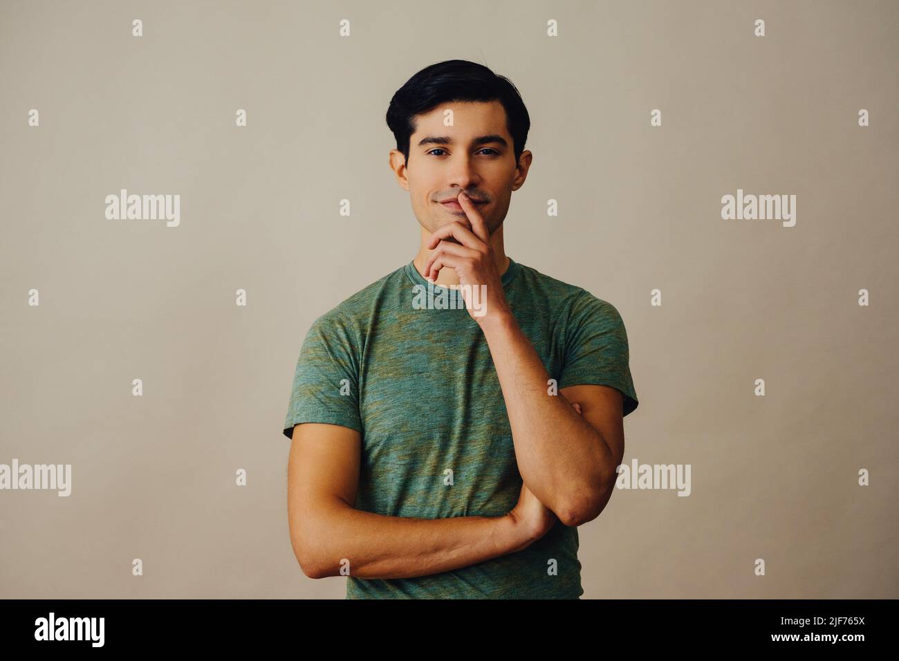 Portrait hispanique latino-homme avec les bras croisés main sur le menton noir cheveux souriant beau jeune adulte vert t-shirt sur fond gris regardant la prise de vue en studio Banque D'Images