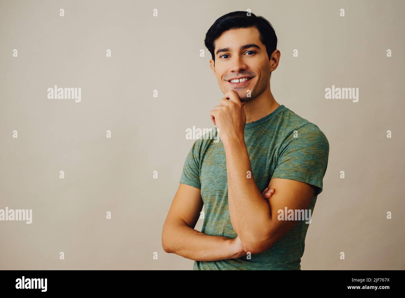 Portrait hispanique latino-homme avec les bras croisés main sur le menton noir cheveux souriant beau jeune adulte vert t-shirt sur fond gris regardant la prise de vue en studio Banque D'Images