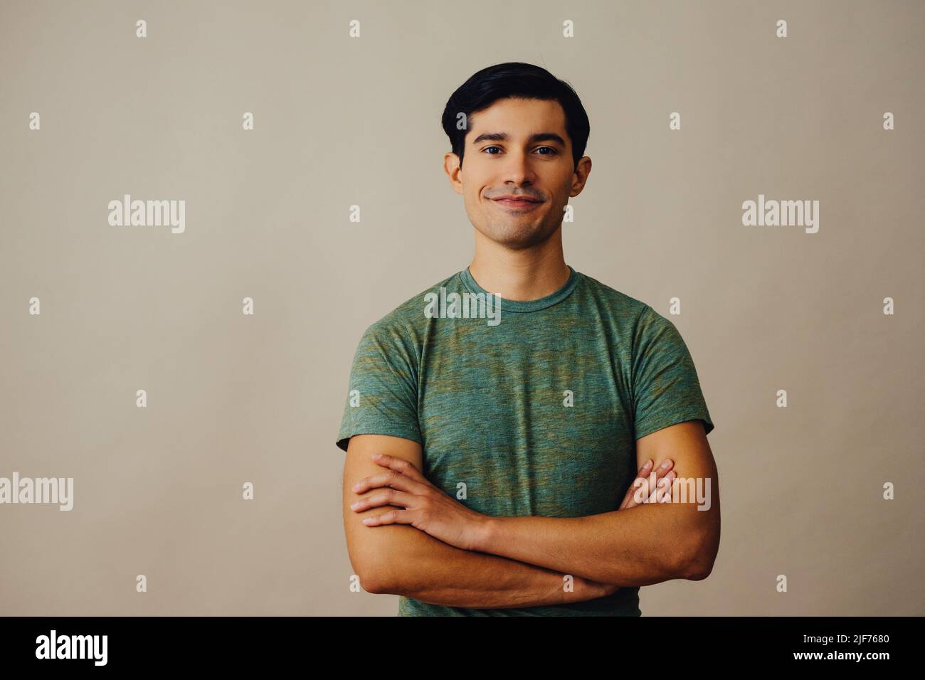 Portrait latino homme avec les bras croisés et les cheveux noirs souriant beau jeune adulte vert t-shirt sur fond gris regardant l'appareil photo en studio Banque D'Images