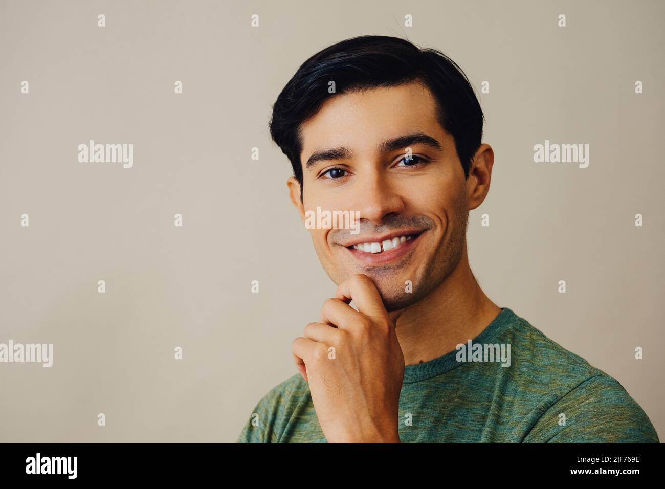 Tête souriante beau jeune adulte latino homme avec main sur menton cheveux noirs et chemise verte sur fond gris regardant la prise de vue en studio Banque D'Images