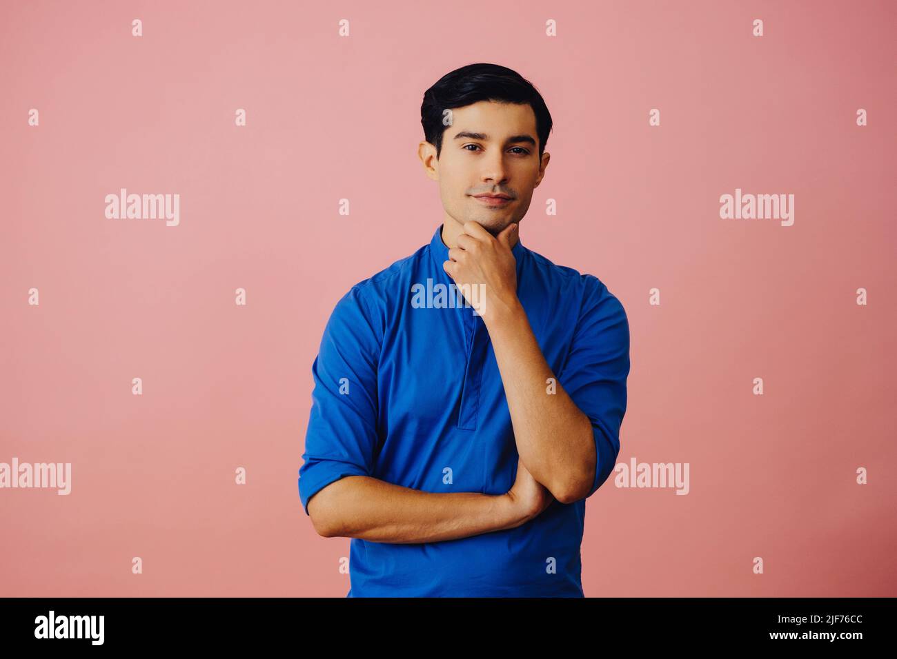Portrait hispanique latino homme avec les bras croisés main sur menton noir cheveux souriant beau jeune adulte bleu chemise sur fond rose regardant la prise de vue en studio Banque D'Images