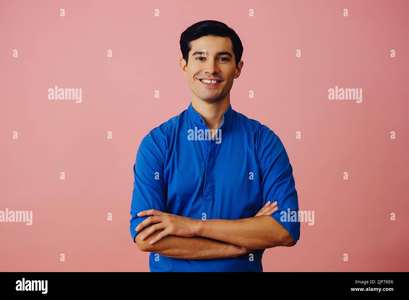 Portrait latino homme avec les bras croisés et les cheveux noirs souriant jeune adulte beau maillot bleu sur fond rose regardant la prise de vue en studio Banque D'Images
