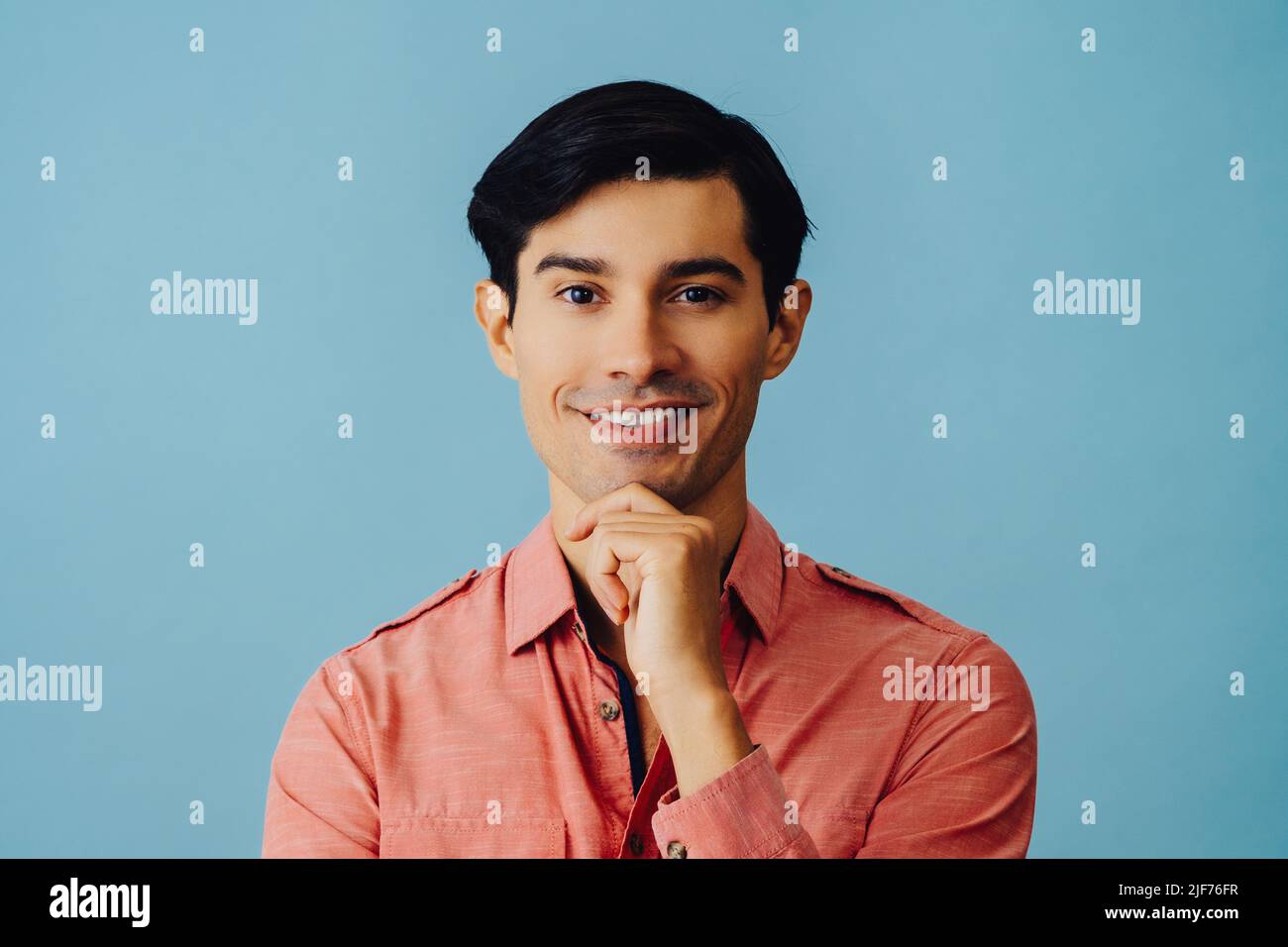 Tête souriante beau jeune adulte latino homme avec main sur menton cheveux noirs et chemise rose sur fond bleu regardant la prise de vue en studio Banque D'Images