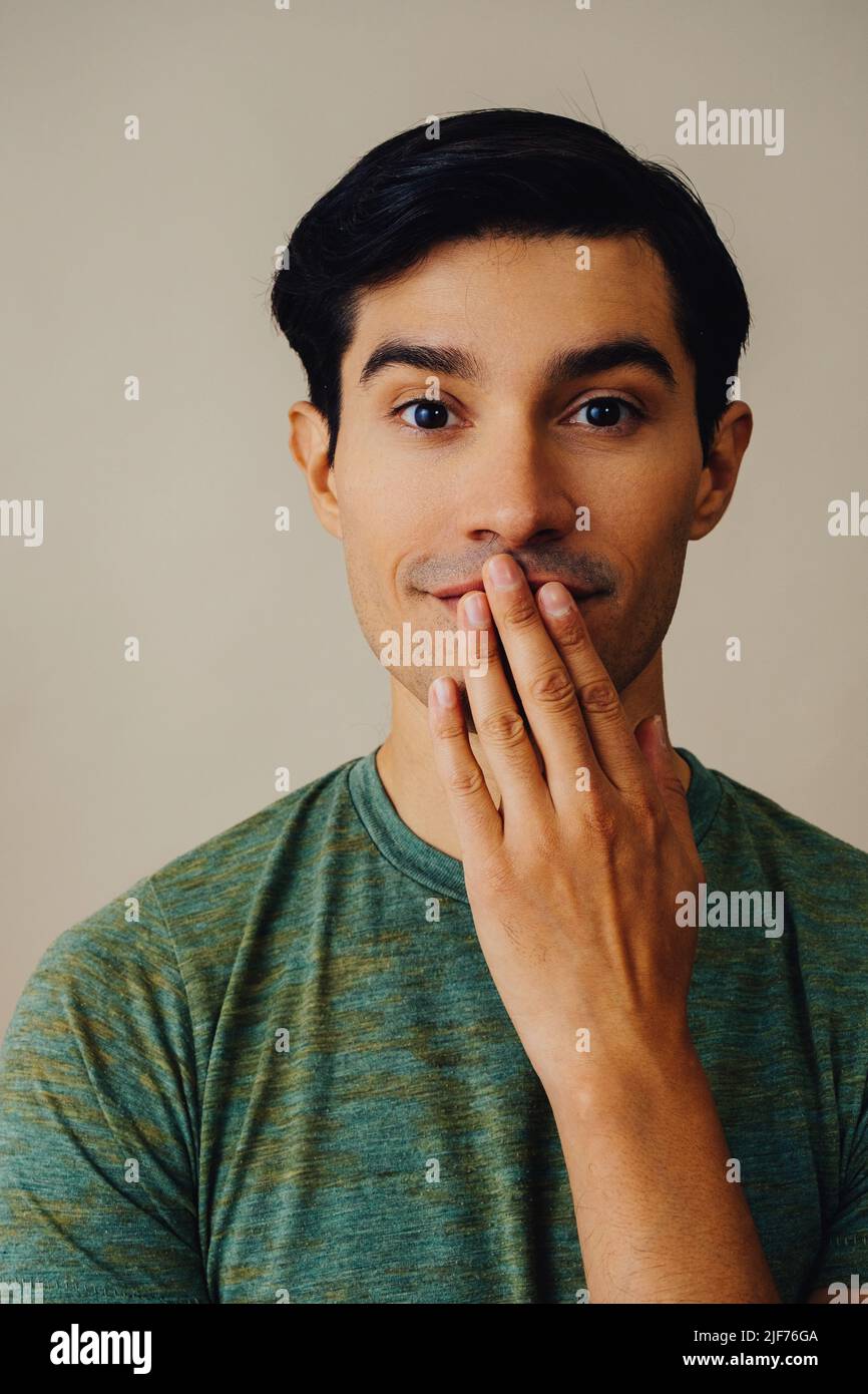 Headshot latino homme noir cheveux main couvrant la bouche beau jeune adulte vert t-shirt sur fond gris regardant la prise de vue en studio Banque D'Images