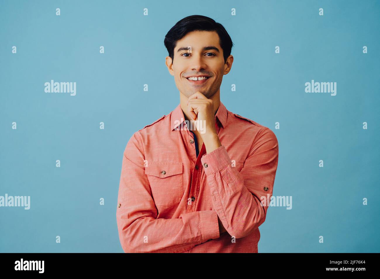 Portrait souriant beau jeune adulte latino homme avec les bras croisés et la main sur le menton cheveux noirs et chemise rose sur fond bleu regardant la photo en studio Banque D'Images
