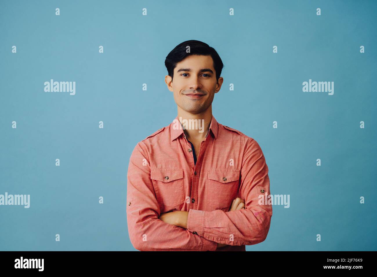 Portrait souriant beau jeune adulte latino homme avec les bras croisés et la main sur le menton cheveux noirs et chemise rose sur fond bleu regardant la photo en studio Banque D'Images