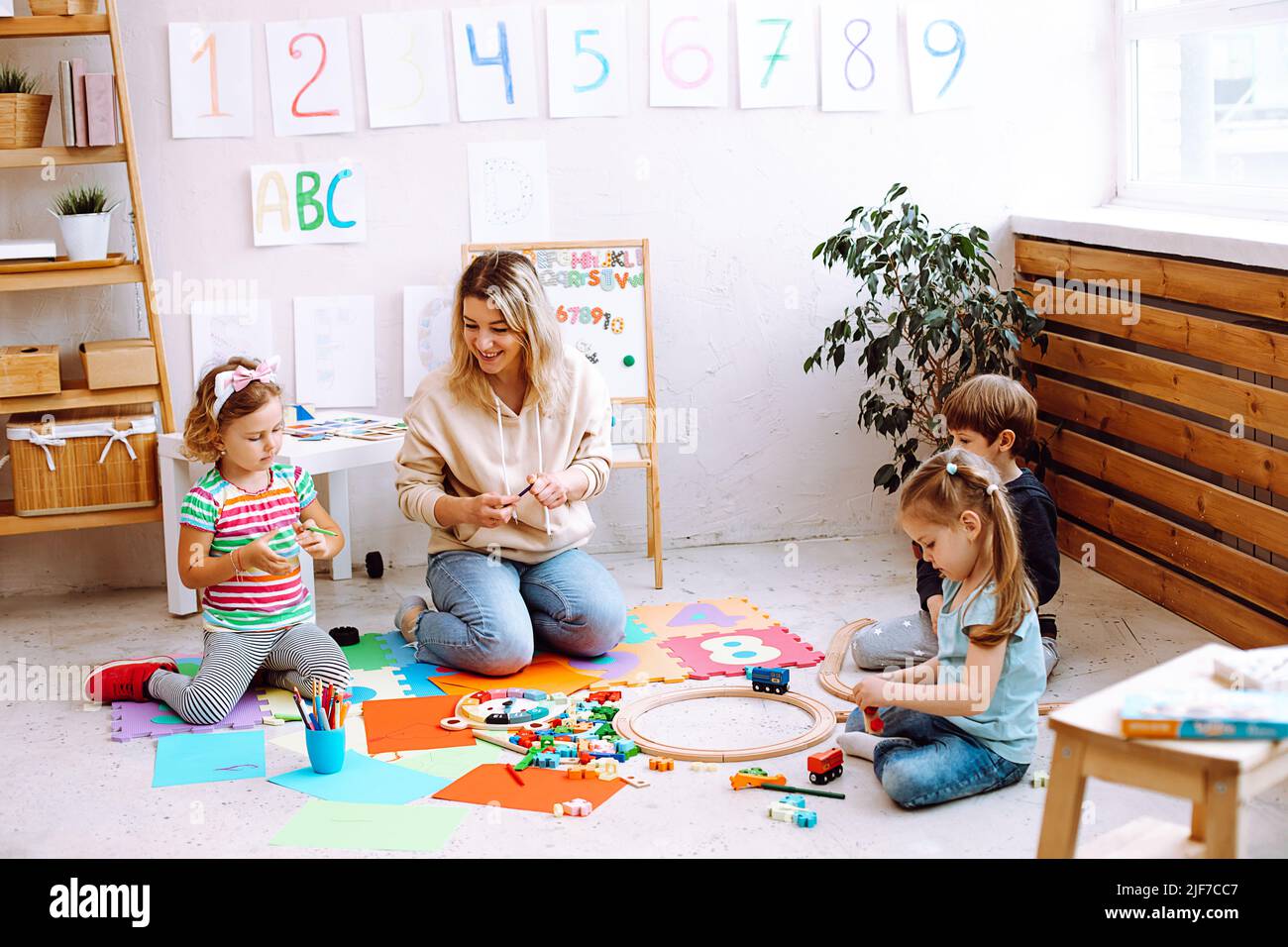Une femme éducatrice joue avec des enfants assis sur le sol à la maternelle. Jouets pliants LEGO pour enfants et dessin avec crayons de couleur dans la salle de jeux. Jouets pour Banque D'Images
