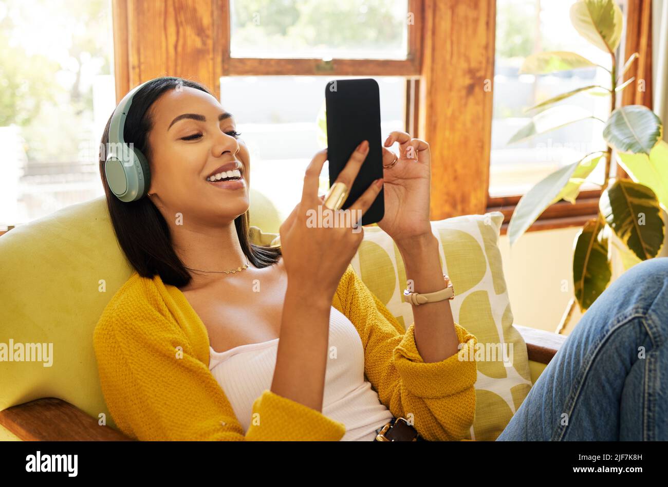 Une femme hispanique utilise son smartphone et écoute de la musique sur un casque tout en étant confortable et relaxante dans un salon lumineux. Une jeune femme Banque D'Images