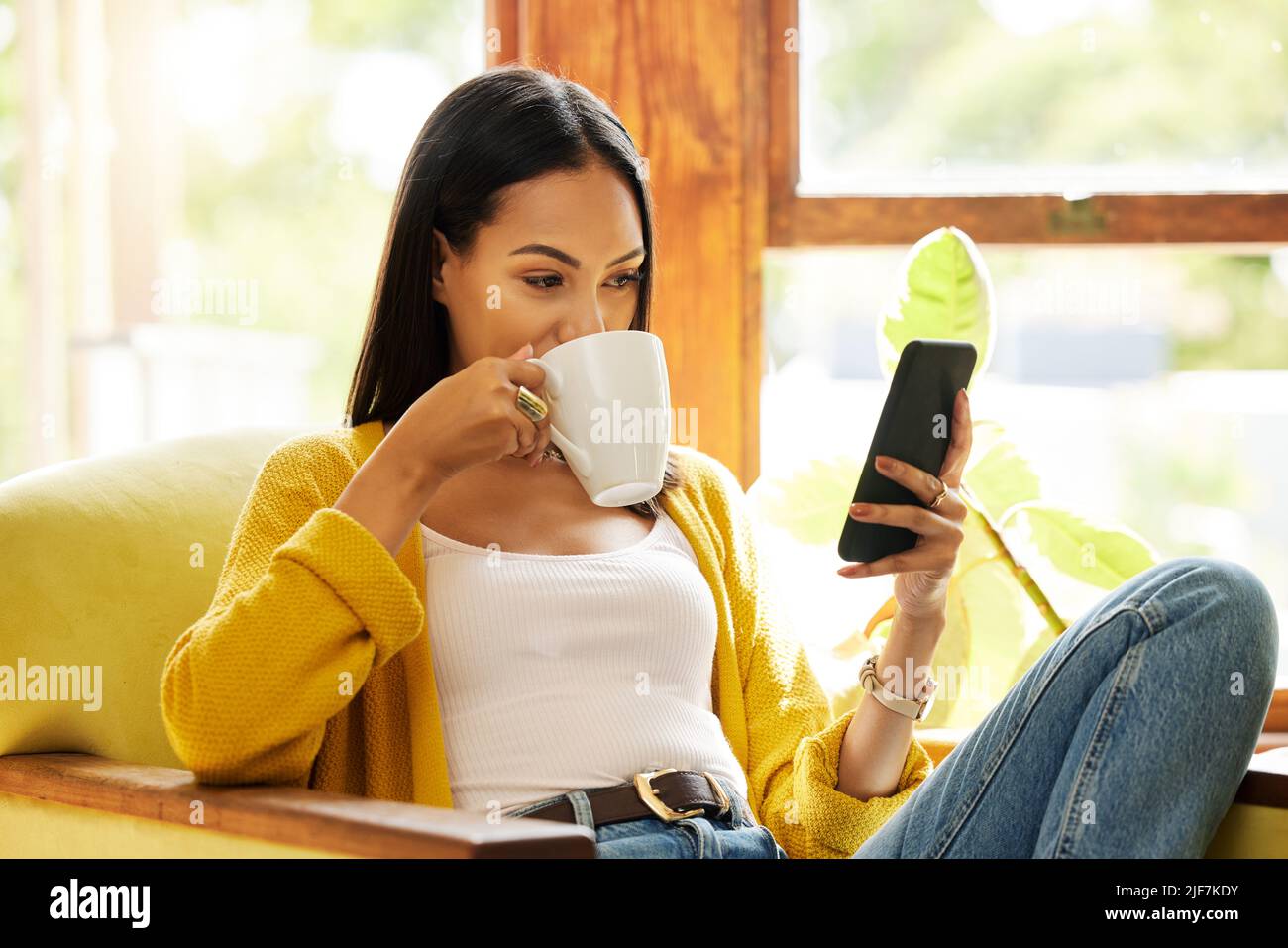 Femme utilisant son smartphone et buvant du café assis devant une fenêtre dans un salon lumineux. Une jeune femme regardant son téléphone portable se détendre Banque D'Images