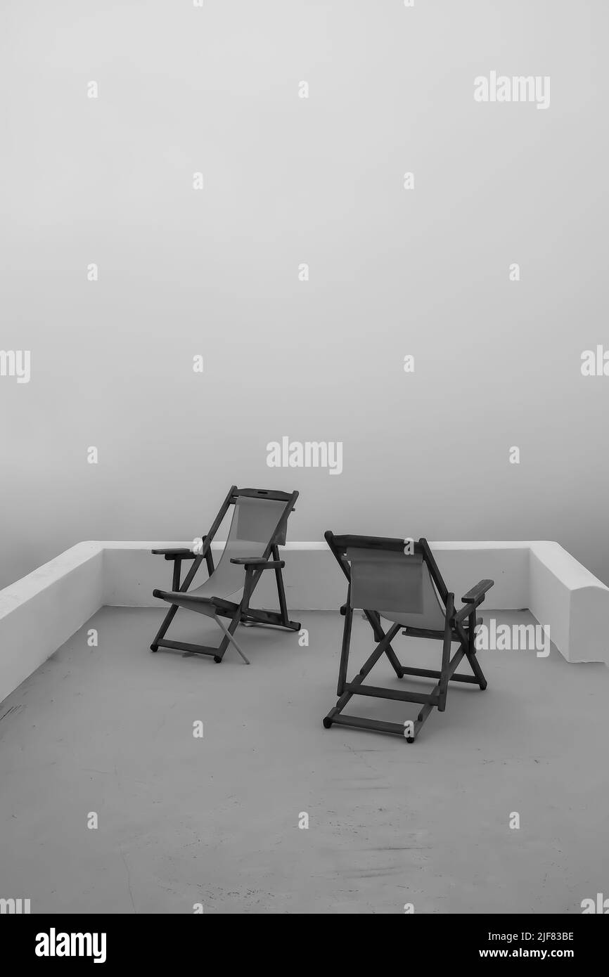 Vue sur deux chaises longues vides en face de la mer égée qui est couverte de brouillard à Santorini Grèce en noir et blanc Banque D'Images