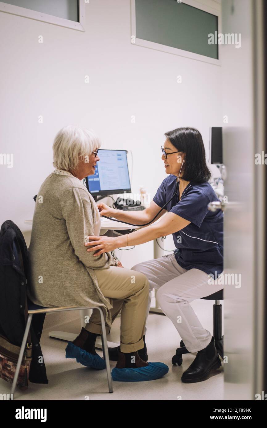 Pleine longueur d'expert en soins de santé féminin avec stéthoscope examinant le patient senior vu par la porte dans la clinique médicale Banque D'Images