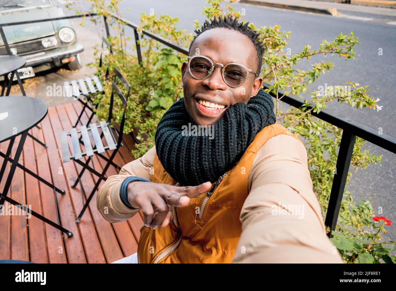 jeune homme africain à l'extérieur, assis à l'extérieur du café sourire heureux prend selfie faisant v signe avec la main, regardant l'appareil photo, perspective du téléphone. Banque D'Images