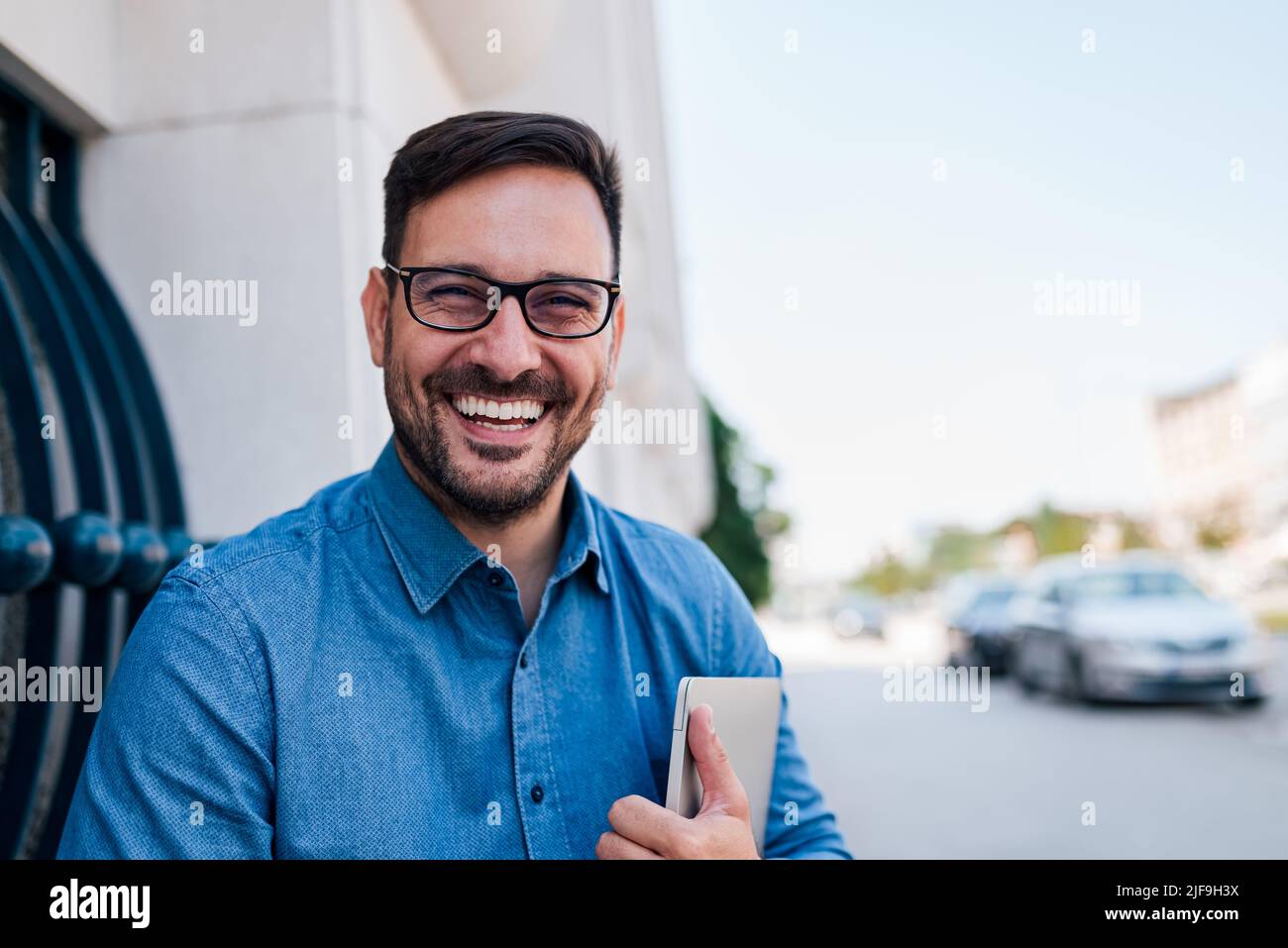 Portrait d'un homme d'affaires souriant tenant un ordinateur portable. Un homme joyeux, un homme de la direction, est assis contre le bâtiment. Il se déplace dans la ville. Banque D'Images