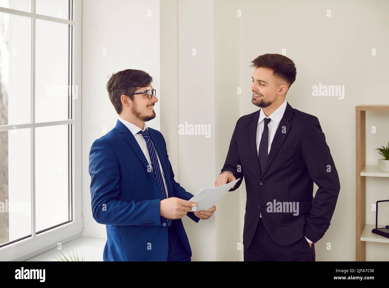 Deux jeunes hommes heureux en costume debout au bureau et discutant de contrat d'affaires Banque D'Images