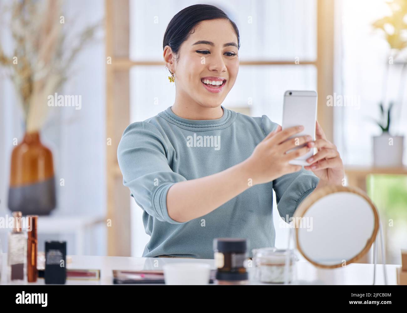 Jeune femme gaie assise à son bureau prenant un selfie sur son téléphone portable avant d'appliquer des produits de soin de la peau. Femme souriante utilisant son smartphone pour Banque D'Images
