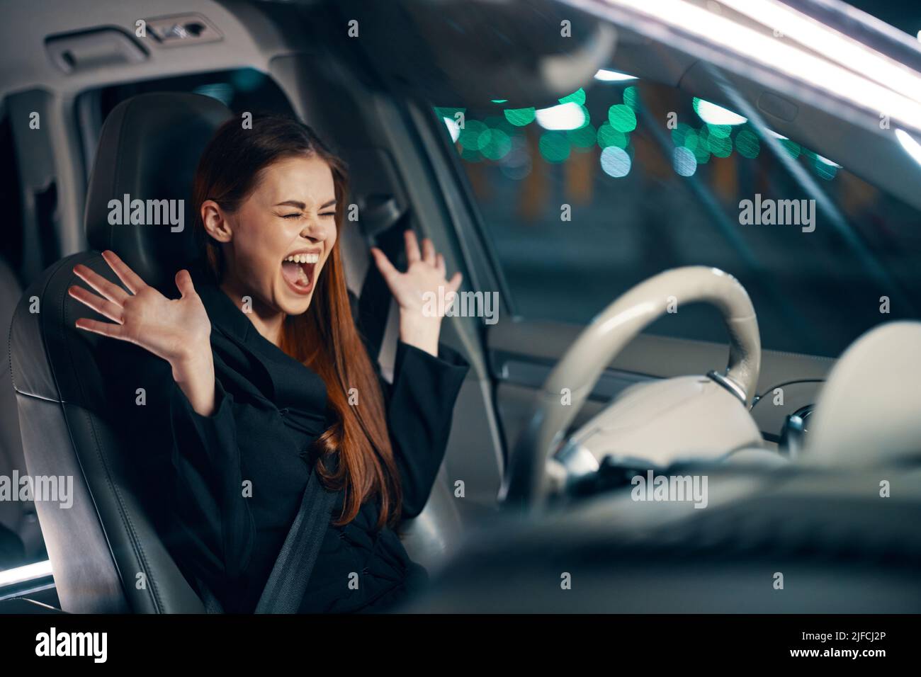 une femme émotif et effrayée est assise derrière le volant d'une voiture dans une chemise noire, portant une ceinture de sécurité, exprimant ses émotions, écartant ses bras Banque D'Images