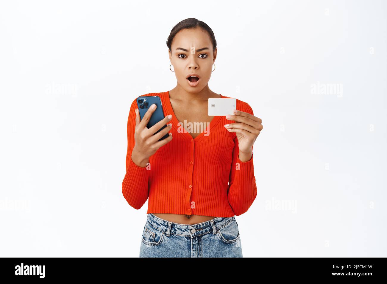 Femme choquée vérifiant téléphone mobile et carte de crédit, regardant inquiet et inquiet, debout sur fond blanc Banque D'Images