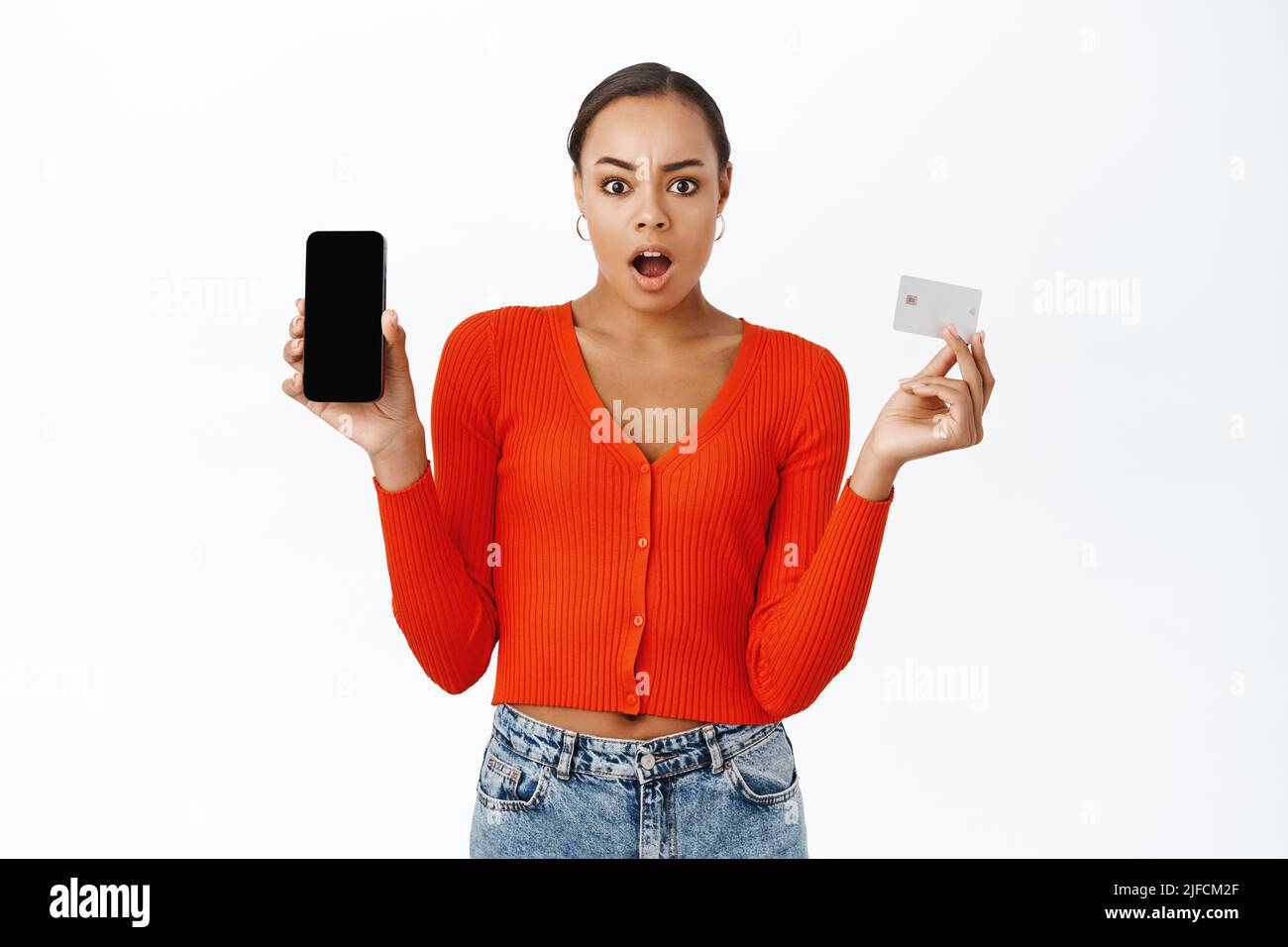 Une femme frustrée montre son téléphone mobile et sa carte de crédit, semble choquée, en attente d'explication, debout sur fond blanc Banque D'Images
