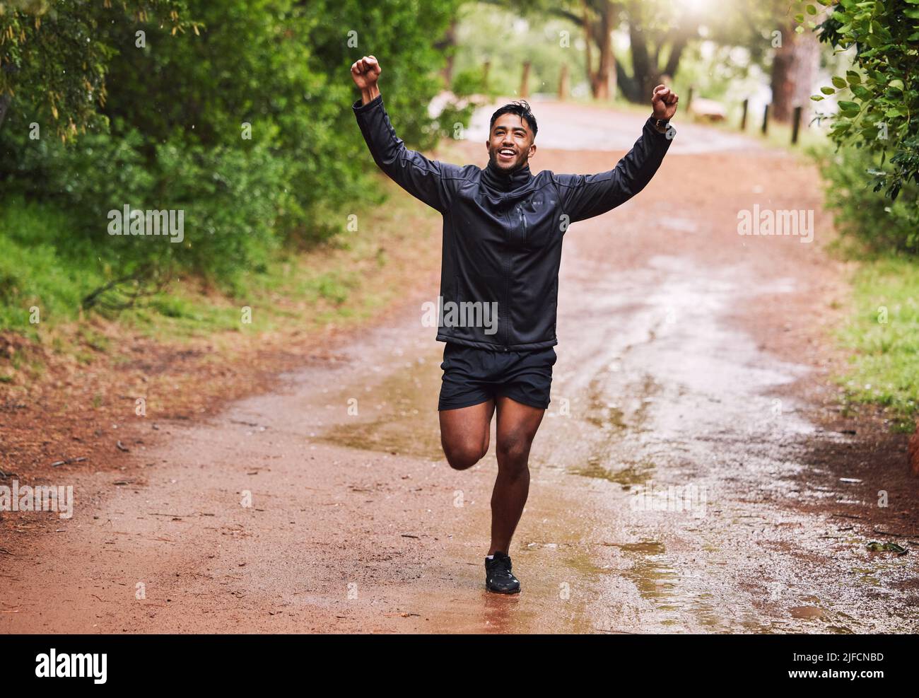 Un jeune athlète hispano-masculin en forme de femme applaudit avec ses poings dans les airs lors d'une course dans une forêt sous la pluie à l'extérieur dans la nature. L'exercice est bon pour Banque D'Images