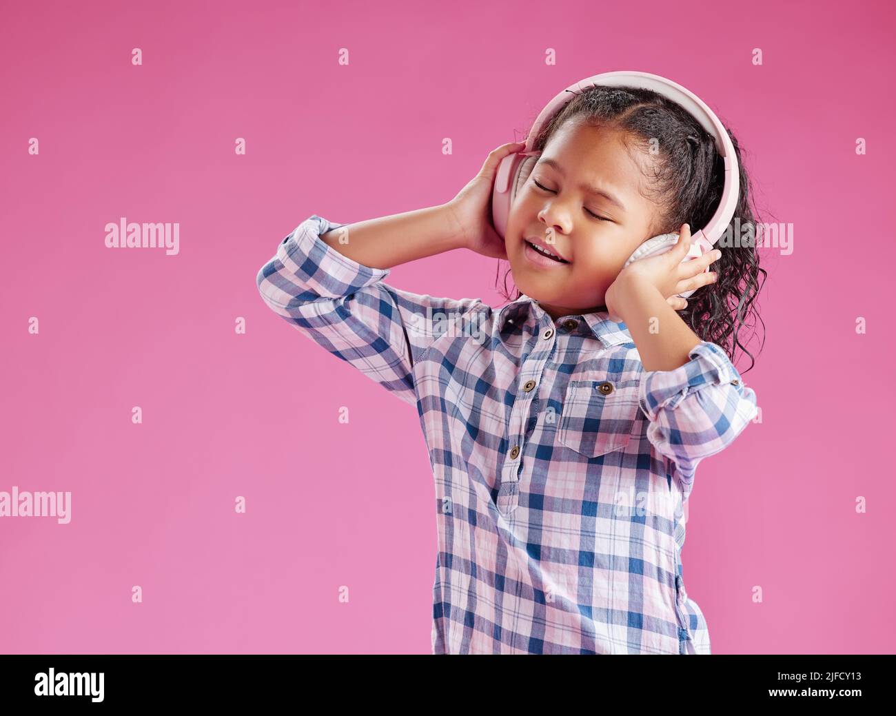 Une jolie petite fille de race mixte avec des cheveux bouclés à l'écoute de la musique tout en portant des écouteurs contre un arrière-plan rose copyspace dans un studio. Africain Banque D'Images