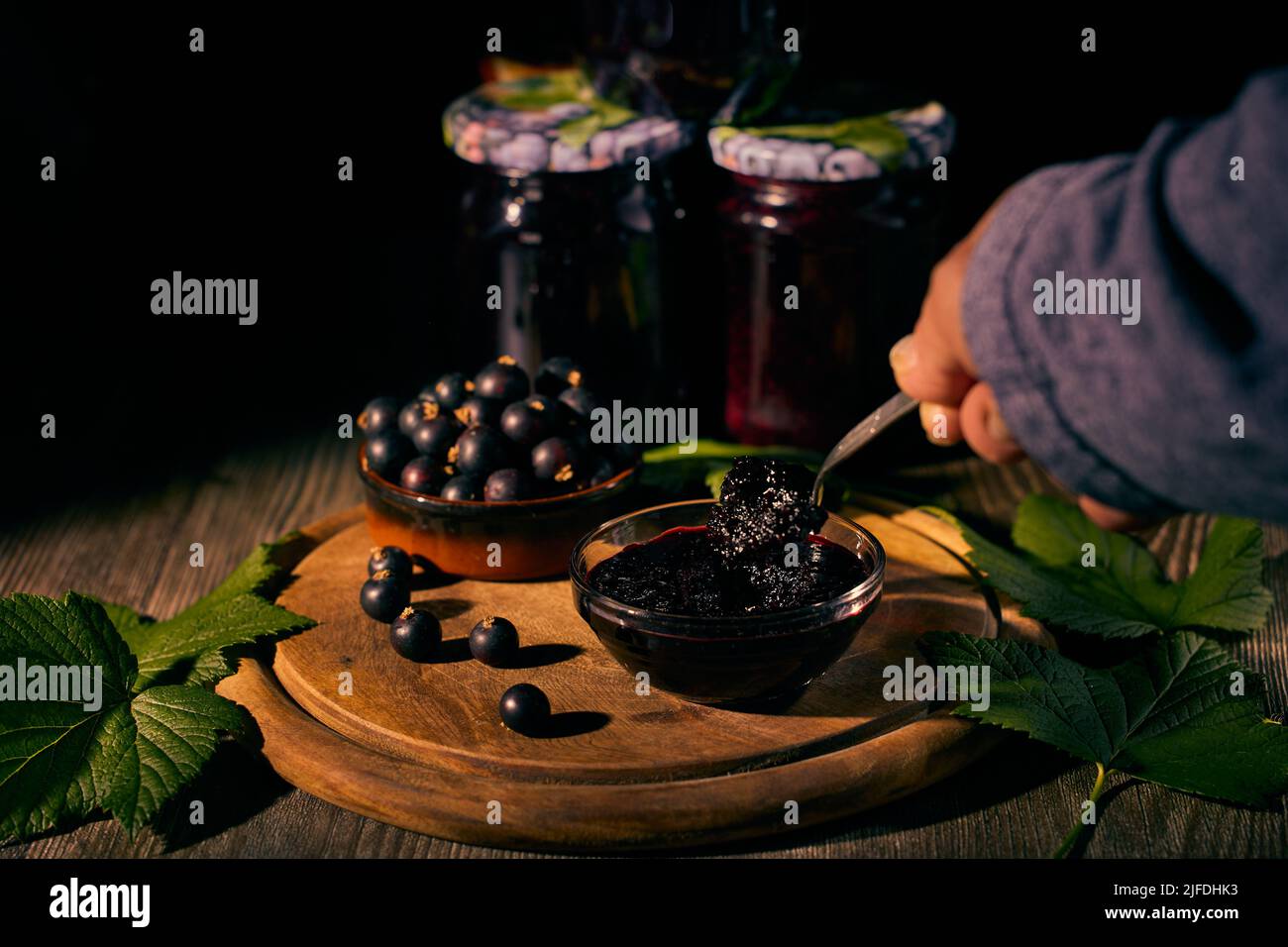 Confiture de cassis dans un bol en verre et cassis dans un bol en céramique sur une planche en bois avec des feuilles de cassis, une confiture de cueillette à la main et des pots en verre de Banque D'Images