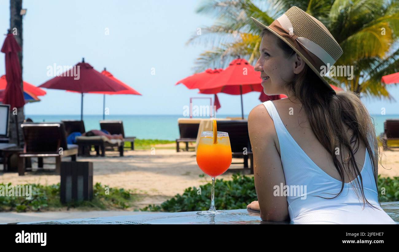 Jeune belle femme dans la piscine en chapeau de paille et maillot de bain, célébrer des vacances dans le pays chaud avec un verre de cocktail d'orange. Été tropical Banque D'Images