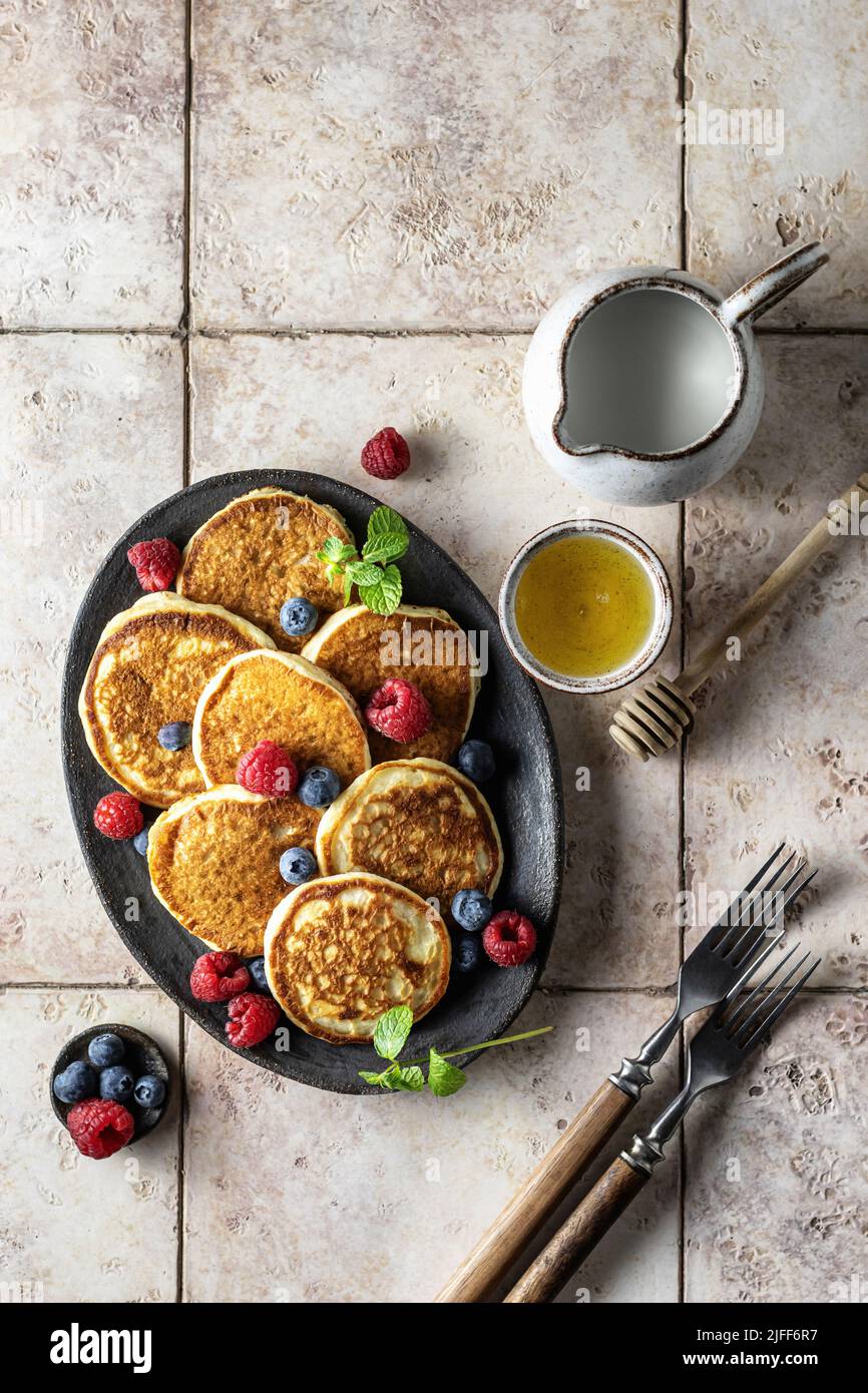 Crêpes aux framboises et aux myrtilles, feuilles de menthe, miel et couverts rustiques sur fond de carreaux roses, vue du dessus Banque D'Images