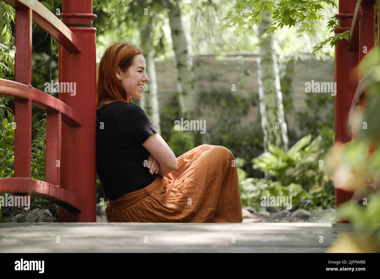 Femme dans son 40s assis sur le sol regardant loin dans le jardin vert. Se reposer, penser. Femelle avec des cheveux de gingembre de l'arrière sur le pont Banque D'Images