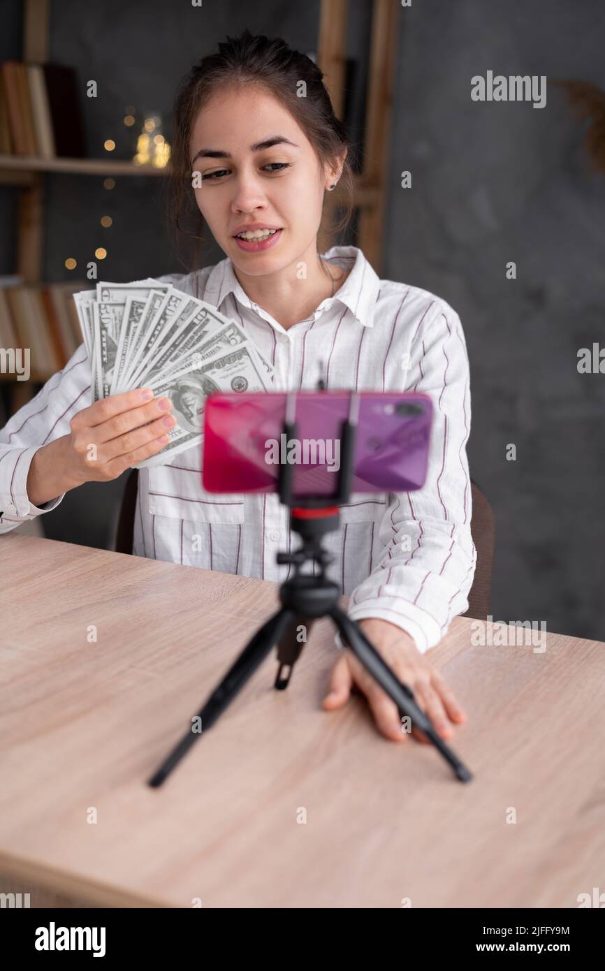 Une jeune femme heureuse avec une banque smartphone facture des billets de banque à partir de l'écran sur fond gris de bureau mural. Technologie virement bancaire en ligne, e Banque D'Images