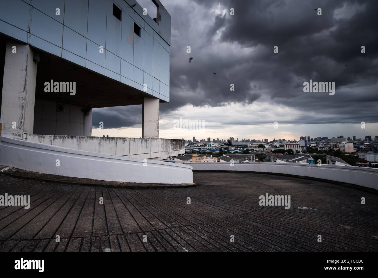 Vue en perspective d'un parking vide sur le toit d'un centre commercial abandonné, des nuages de pluie sombres couvrent une ville en arrière-plan. Banque D'Images