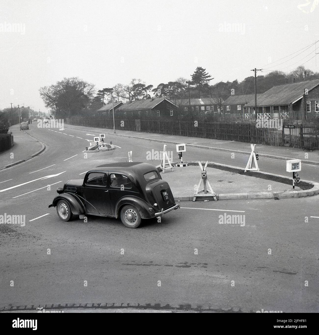 1960s, historique, une voiture Ford Anglia de l'époque à une jonction de route triangulaire nouvellement érigée sur Slade Rd, Oxford, Angleterre, Royaume-Uni. L'image de droite montre les anciens camps de l'armée au large de la Slade, qui ont fourni des logements très nécessaires aux familles dans la période d'après-guerre. Banque D'Images