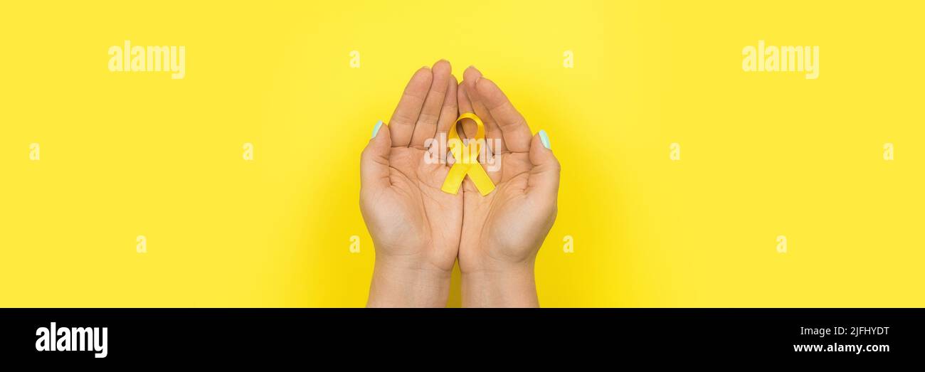 Ruban d'or jaune dans les mains d'une fille sur fond jaune. Concept de cancer pour enfants. Mois de sensibilisation. Journée du cancer chez l'enfant février 15. Web Banque D'Images