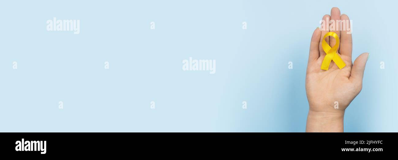 Ruban d'or jaune dans la main d'une femme sur fond bleu. Concept de cancer pour enfants. Mois de sensibilisation. Journée du cancer chez l'enfant février 15. Web Banque D'Images