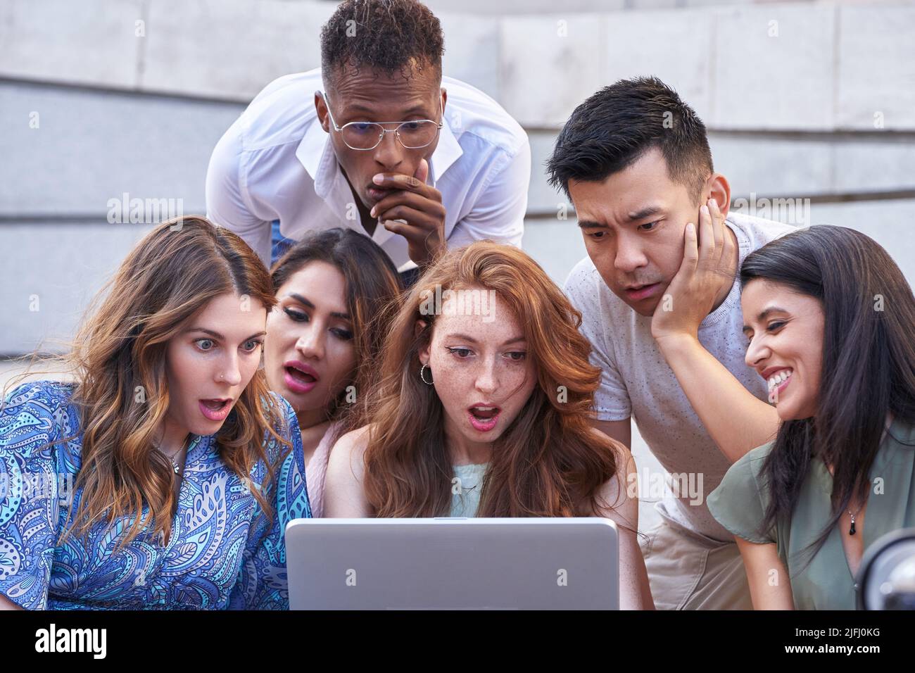 Groupe de personnes multiethniques regardent l'écran d'ordinateur portable avec une expression choquée sur leurs visages tout en étant assis sur les escaliers à l'extérieur. Mode de vie urbain, technologie et concept d'amitié. Banque D'Images