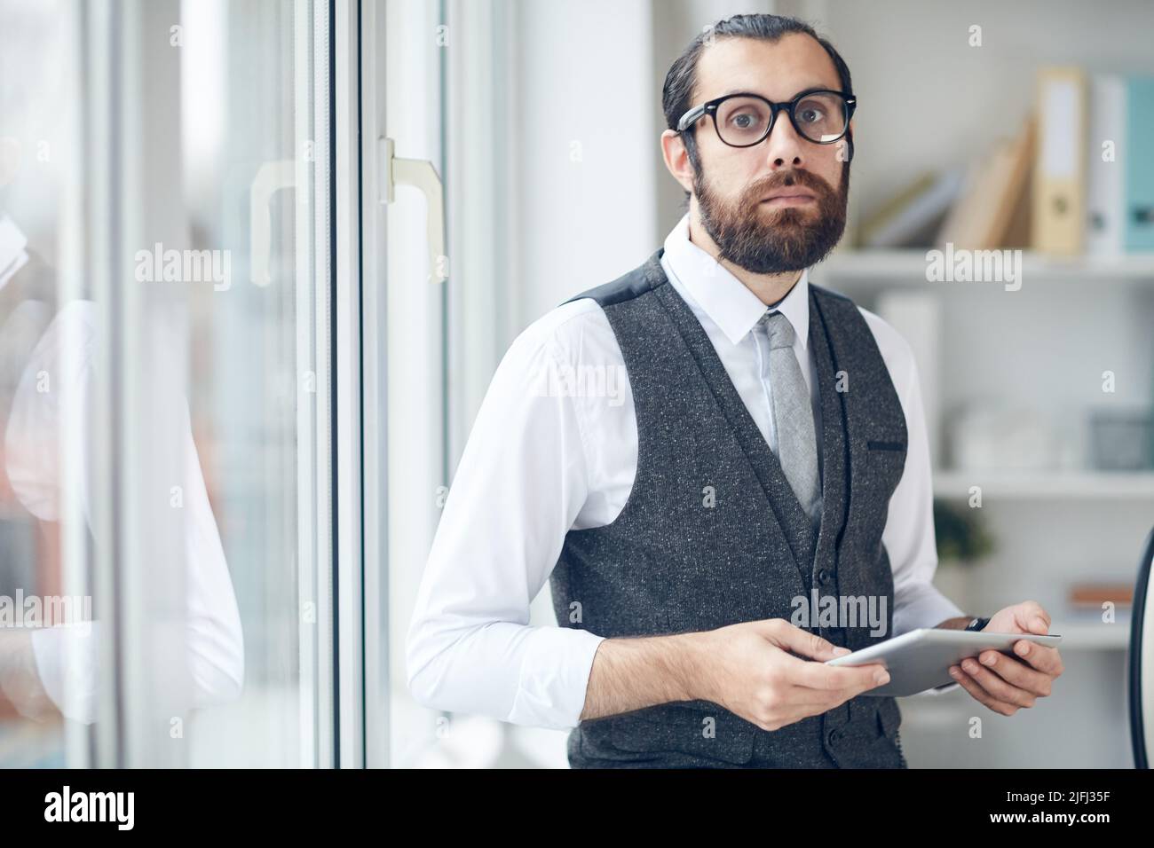 Portrait d'un triste directeur d'entreprise nerd en gilet de costume et chemise blanche debout au bureau et utilisant une tablette numérique Banque D'Images